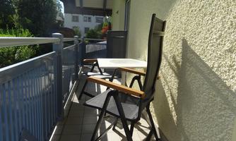 A small balcony with two chairs and a table. The sun is shining and there is plenty of green foliage in the background.