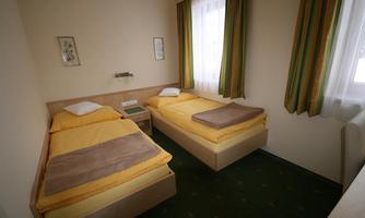 A simple room with two single beds, yellow bedspreads, and a small nightstand. The walls are light, and there are large windows with green curtains.