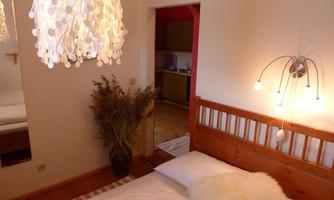 A cozy bedroom with a wooden bed and a striking lamp. In the background, a small kitchen and decorative plants can be seen.
