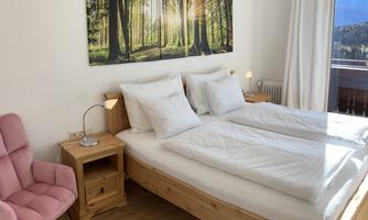 A cozy bedroom with a double bed and wooden furniture. On the wall hangs a picture of a forest area bathed in soft light.