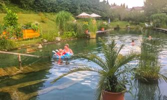 A quiet pond surrounded by greenery with bathers. Two children are playing in the water while others relax.