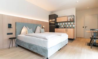 A modern room with a large double bed and a small kitchenette. The space has a bright, friendly atmosphere and stylish furniture.