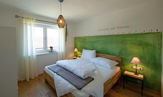 A cozy bedroom with a wooden bed and white bedding. There is a green accent on the wall, and there is a window with curtains.