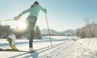A person is cross-country skiing on a snowy trail. In the background, there are mountains and a clear blue sky.