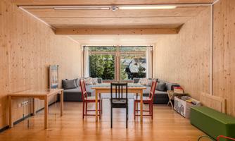 A bright room with wooden paneling and large windows. There are tables, chairs, and a cozy seating area in the room.