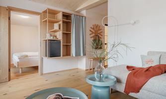 A modern living room with a cozy sofa and a small table. In the background, you can see a bedroom with a large bed.