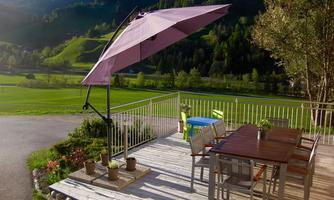 A cozy outdoor area with a wooden terrace table and a large sun umbrella. In the background, green hills and trees can be seen.