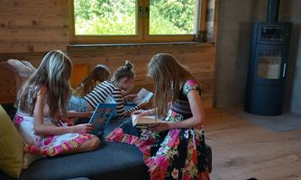 Four girls are sitting on a sofa reading books. The room is bright with large windows and a modern fireplace.