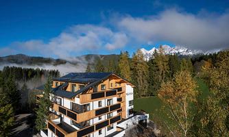 A modern building surrounded by trees and mountains. The sky is clear with some clouds.