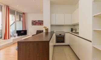 A modern kitchen with white cabinets and a wood workspace. The space is bright and friendly with large windows and a cozy seating area.