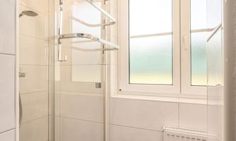A modern bathroom with a glass shower and large windows. The room is bright and clean with white tiles and a dark floor.