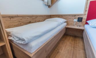 A simple bedroom with a single bed and wooden paneling. The room is bright and furnished with a nightstand.