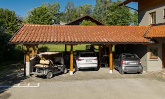 A covered parking area with two cars and a golf cart. In the background, trees and a building are visible.