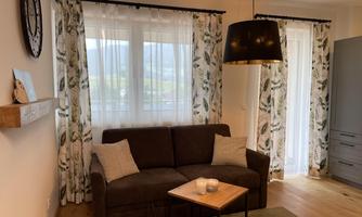 A cozy living space with a brown sofa and a small table. Large windows let in plenty of daylight and the curtains have a floral pattern.