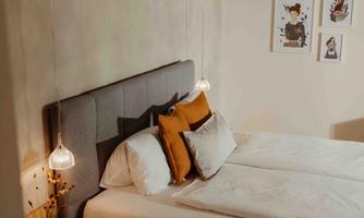 A cozy bedroom with a gray headboard and decorative cushions in warm tones.  
Artworks hang on the wall, and soft lighting creates an inviting atmosphere.