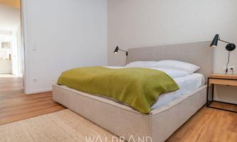 A modern bedroom with a comfortable bed and a green wool blanket. The warm wooden floors and the minimalist lamps create a cozy atmosphere.