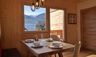 A cozy dining area with a wooden table and a set table. In the background, there is a large window front with a view of the mountains.