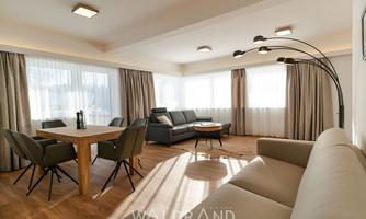A modern living room with large windows and plenty of natural light. There is a cozy seating area and a wooden dining table.