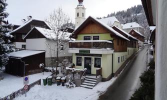 A snowy street in a small village. In the background, a church tower and some classic huts can be seen.
