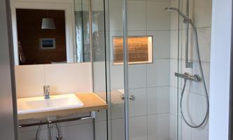 A modern bathroom with a glass shower and a bright sink. The walls are tiled and the floor is made of wood.