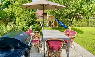 A cozy garden with a table and chairs under a parasol. In the background, there is a playground swing and a slide.