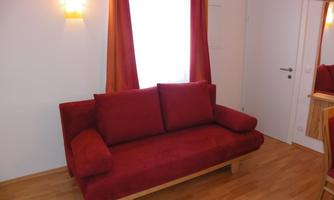 A red sofa with wide armrests is located in a bright room. Next to the sofa, curtains in warm colors and a window are visible.
