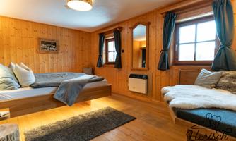 A cozy bedroom with wooden walls and large windows. There is a double bed, a seating area, and a warm atmosphere.