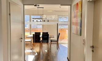 A bright hallway with wooden flooring leading to a dining area. In the background, a table and chairs as well as a large window area are visible.