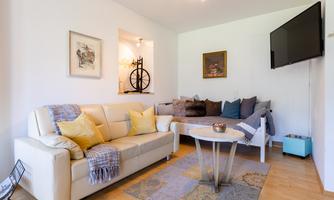 A cozy living room with a beige sofa, cushions, and a small table. Pictures hang on the wall, and there is a TV as well as a bright atmosphere.