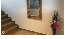 A bright corridor with a wooden staircase and a decorative mirror on the wall. Next to the mirror is a flower arrangement.