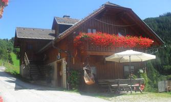 A traditional wooden house with red flower boxes. Outside, there is a table with an umbrella in a quiet, mountainous setting.