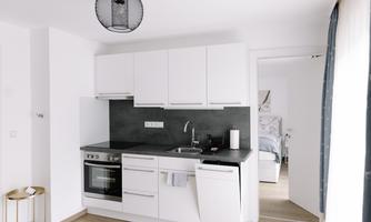 A modern, open kitchen with white cabinets and a dark countertop. In the background, a bright living area is visible.