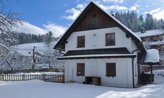 A cozy house in winter, surrounded by snow-covered trees. The sky is clear and blue, creating a tranquil atmosphere.