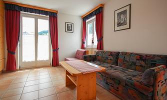 A cozy living room with a colorful sofa and a wooden table. Large windows with red curtains let in a lot of light.
