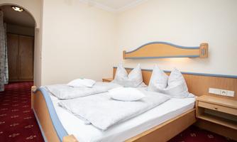 A cozy bedroom with a double bed and several pillows. The walls are painted in neutral colors, and the carpet has a red pattern.