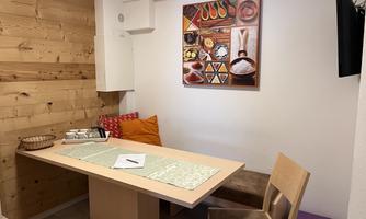 A cozy dining area with a wooden table and chairs. A colorful wall tapestry with various foods hangs on the wall.