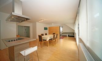A modern living room with a dining area and an open kitchen. The space is bright and friendly, with large windows and minimalist design.