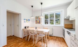 A modern kitchen with a dining table and chairs. Large windows let in plenty of daylight.