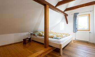 A bright bedroom with a double bed and wooden beams. The window brings daylight into the room, which creates a cozy atmosphere.