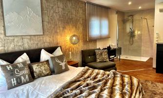 A modern bedroom with a large bed and cozy pillows. In the background, there is a bathroom with a glass shower.