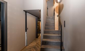 A bright hallway with a wooden staircase. On the right side, there is a smooth wall with a modern sliding door.