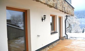 A modern house with a stone facade and large windows. The wooden terrace offers a view of a snow-covered landscape.
