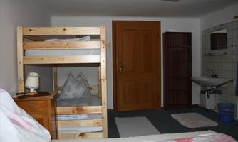A simple bedroom with a bunk bed and a single bed. The room also features a dresser, a sink, and a wardrobe.
