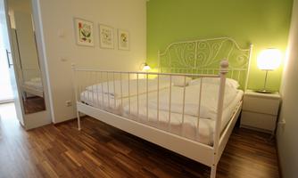 A cozy bedroom with a white bed and a green wall. Next to the bed, there is a bedside lamp and a sideboard.