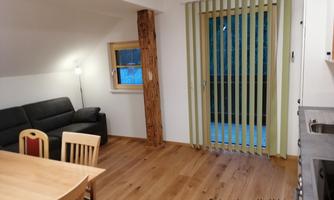 A cozy living area with a black sofa and a dining table. Large windows with blinds provide access to a balcony.