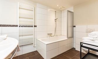 A modern bathroom with a bathtub and a shower. There are light gray tiles and towels on a shelf.