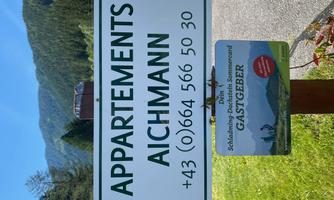 A sign for the Aichmann apartments with contact details. The surroundings are green and mountainous.