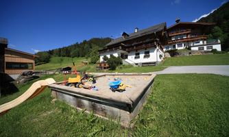 A playground with a sandpit, colorful toys, and a slide. In the background, cozy houses are situated in a green landscape.