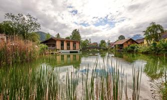 A tranquil pond surrounded by houses and lush vegetation. The sky is cloudy, creating a peaceful atmosphere.