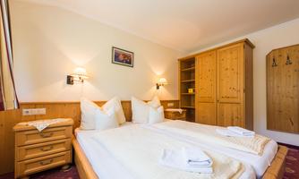 A cozy bedroom with a double bed and bright wooden furniture. The cozy atmosphere is enhanced by the warm wall lamps and the red carpet.